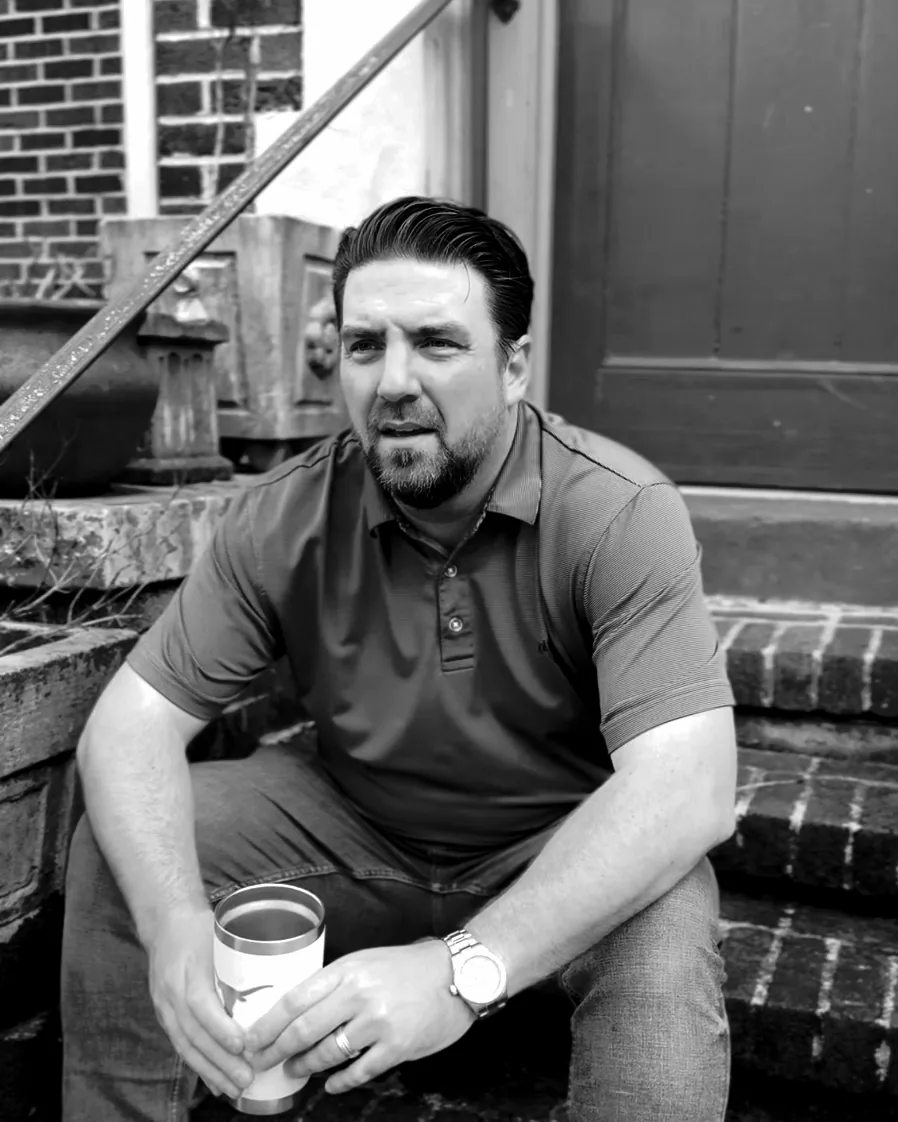 Dustin Weaver sitting on old brick steps by a green door, coffee in hand, mid-thought.